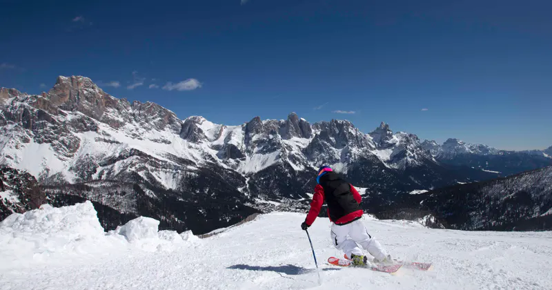Een skiër in een rode jas en een witte broek skiet van een besneeuwde berghelling af, omringd door met sneeuw bedekte bergtoppen onder een helderblauwe hemel.